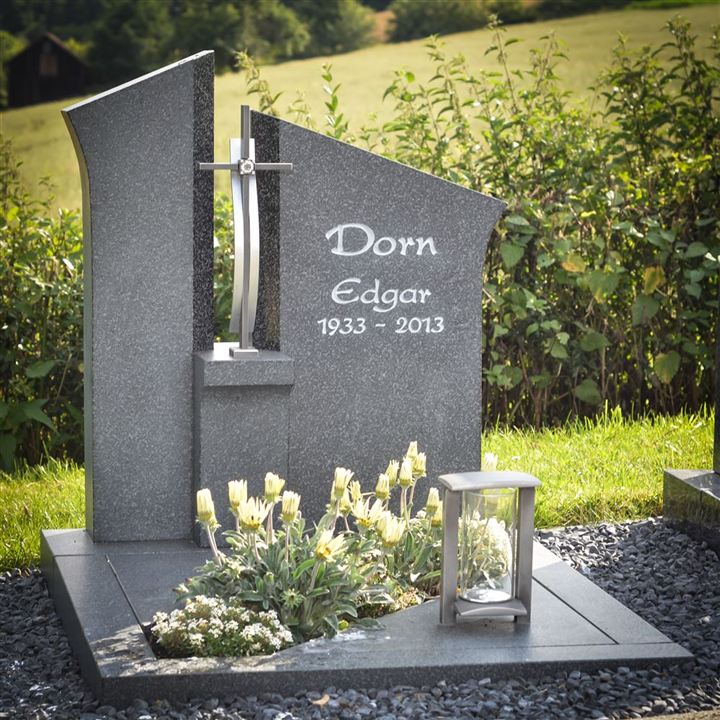 Ein Grabmal aus grauem Naturstein mit einem silbernen Kreuz in einer geometrischen Form mit einem silbernen Kreuz vor einem freien Bereich